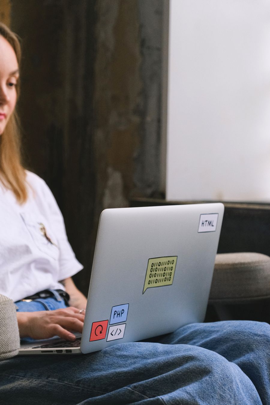 A woman in a white T-shirt works on a laptop resting on her lap, its lid covered in HTML and PHP stickers