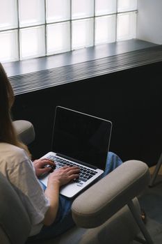 A woman sits on an armchair typing on a laptop in a contemporary office environment.