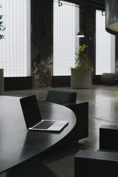 A sleek laptop rests on a black table in a contemporary industrial interior with greenery.