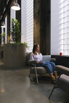 A woman sitting in a stylish office lounge using a laptop, surrounded by natural light and greenery.