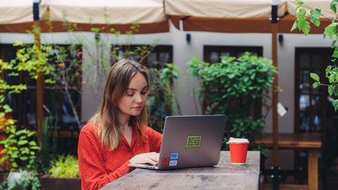 Young woman working on a laptop in an outdoor cafe setting, enjoying a cup of coffee.