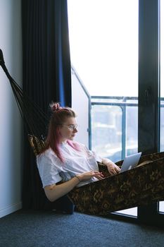 A woman relaxes in a hammock indoors while working on her laptop, embodying the digital nomad lifestyle.