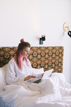 Young woman with pink hair working on a laptop in bed, showcasing a relaxed and productive lifestyle.