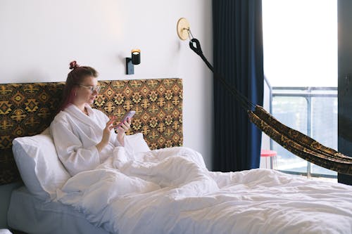 Free A Woman in White Bathrobe Sitting on the Bed while Using Her Phone Stock Photo