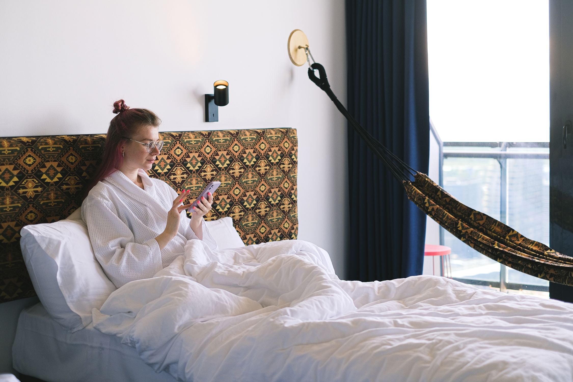 Woman in a bathrobe sits on a bed using a smartphone in a comfortable hotel room.