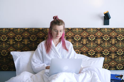 Caucasian woman in robe, working remotely on her laptop from bed, emphasizing comfort and modern technology.