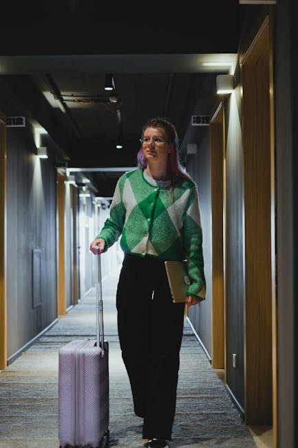 Woman walking through hotel corridor with suitcase, wearing green cardigan.