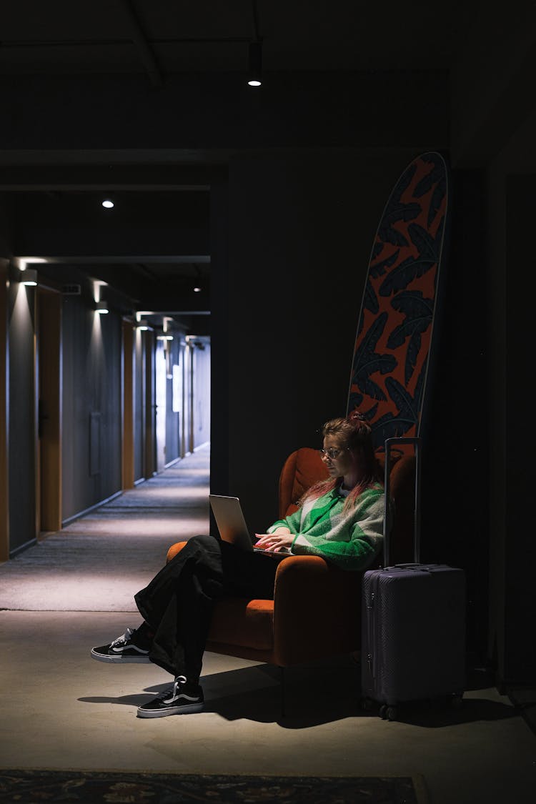 Woman Working On Laptop In Hotel Corridor With Suitcase By Her Side