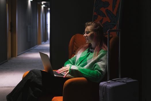 A woman in a hotel hallway using a laptop, sitting on an armchair next to luggage, creating a cozy working environment.