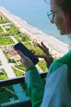 A woman with colored hair uses a smartphone on a balcony overlooking a scenic ocean view.