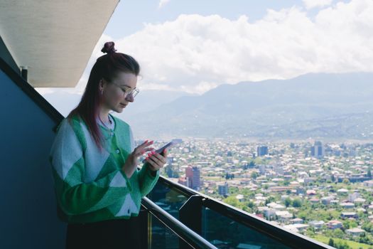 Woman in green sweater using smartphone on a balcony overlooking a cityscape with mountainous backdrop, showcasing digital nomad lifestyle.