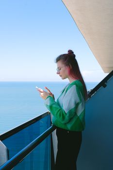Young woman standing on a balcony, using a smartphone with an ocean view. Vibrant and relaxing setting.