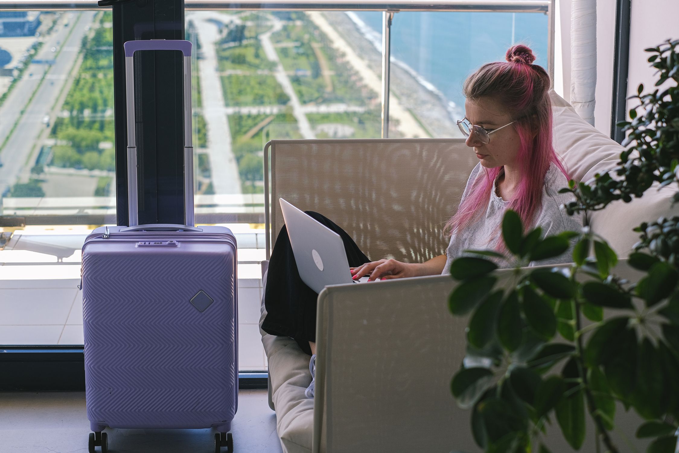 Woman with pink hair working on laptop in hotel room with scenic view out the window