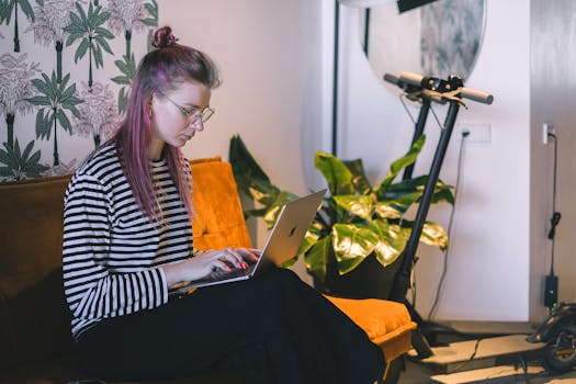 A woman with dyed hair types on a laptop while sitting on a couch in a stylish indoor setting with a scooter nearby.