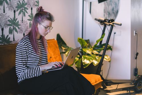 Free A woman with dyed hair types on a laptop while sitting on a couch in a stylish indoor setting with a scooter nearby. Stock Photo