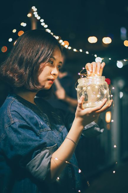 Asian woman holding a glowing jar filled with string lights outdoors at night.