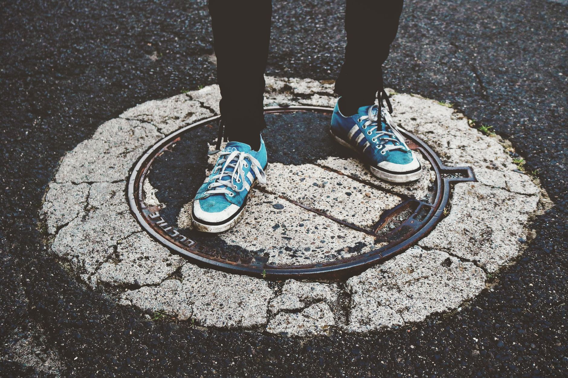 Blue sneakers standing on a weathered manhole cover in a city street.