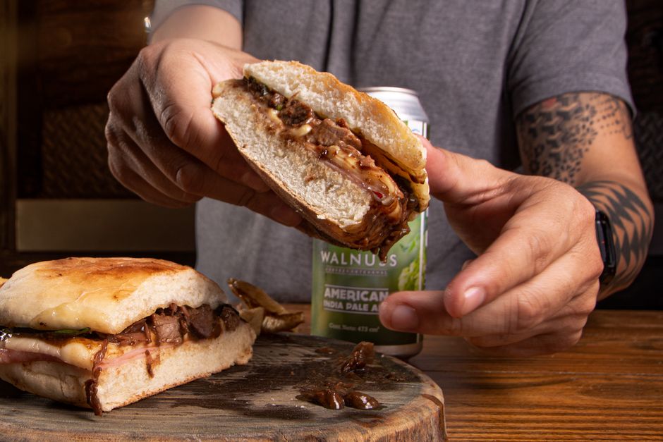 Close-up of hands holding a juicy sandwich with a beer in the background on a wooden board. — Bifana, Portugal.