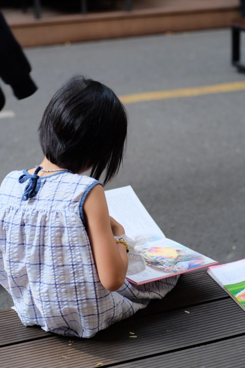 Free Young child in a white dress reading a colorful book while sitting on an outdoor bench. Stock Photo