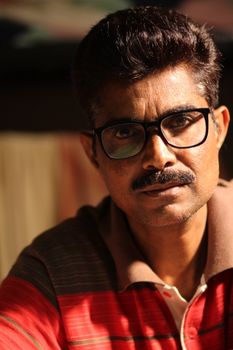 Close-up portrait of a South Asian man with glasses and mustache, lit with warm light.
