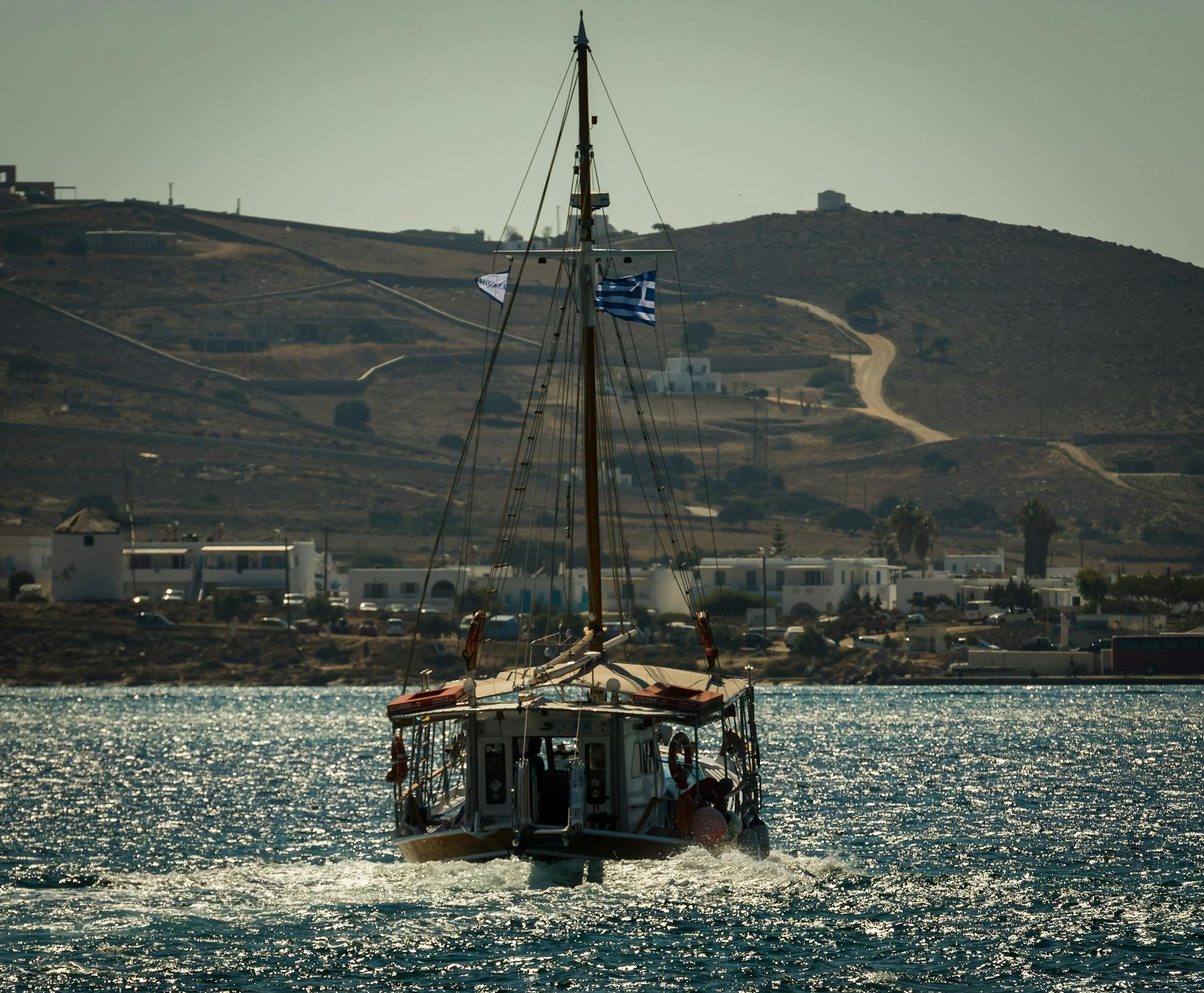A traditional sailing boat with a Greek flag cruising in the Aegean Sea under a clear sky.