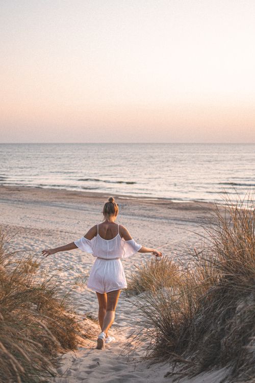 Free A woman in a white outfit walks towards the ocean on a sandy beach during sunset. Stock Photo