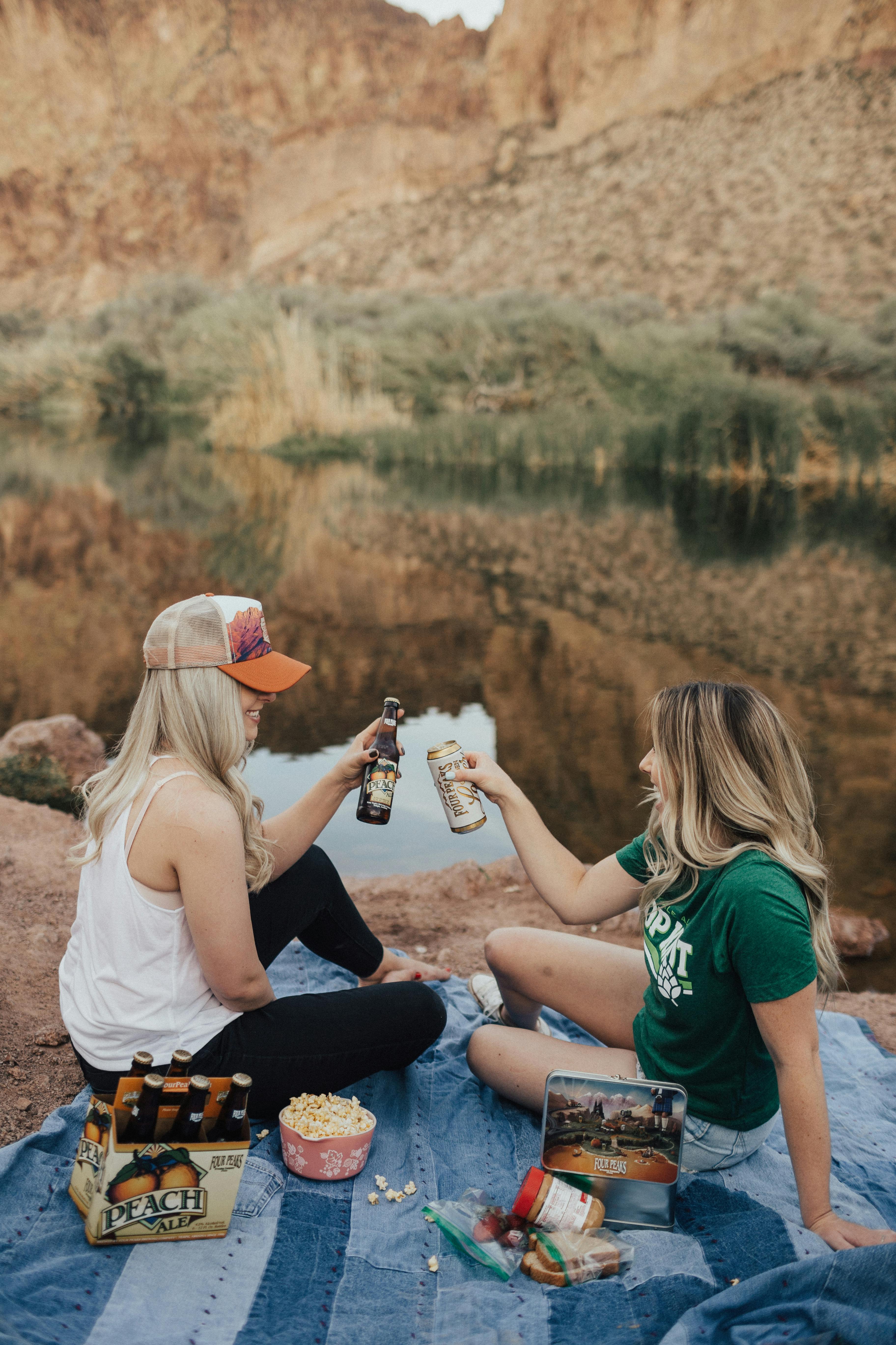 two women holding beer while sitting on rock near body of water