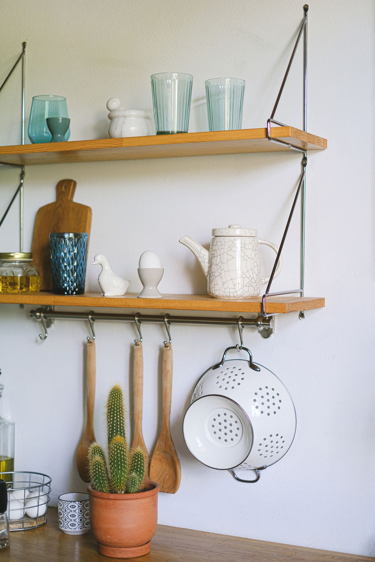 Wooden wall shelves in a kitchen holding glasses and a teapot, with utensils and a colander hanging from hooks below