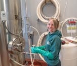 一名穿着绿色夹克、戴着手套的妇女在啤酒厂工作，正站在不锈钢设备旁微笑