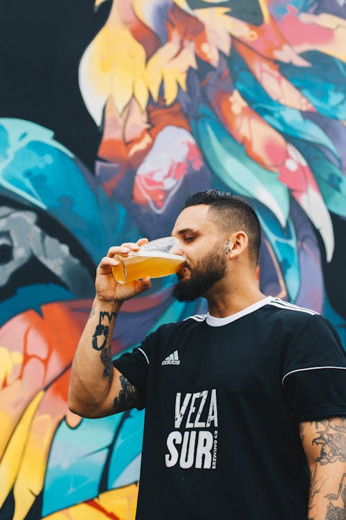 Татуированный мужчина пьет на прозрачном стакане перед разноцветной расписной стеной