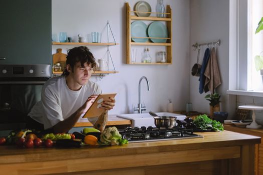 Adult man using a tablet while cooking with fresh vegetables in a modern kitchen setting.