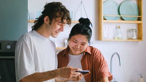 Happy couple using smartphone and preparing vegetables in modern kitchen. A moment of shared culinary joy.