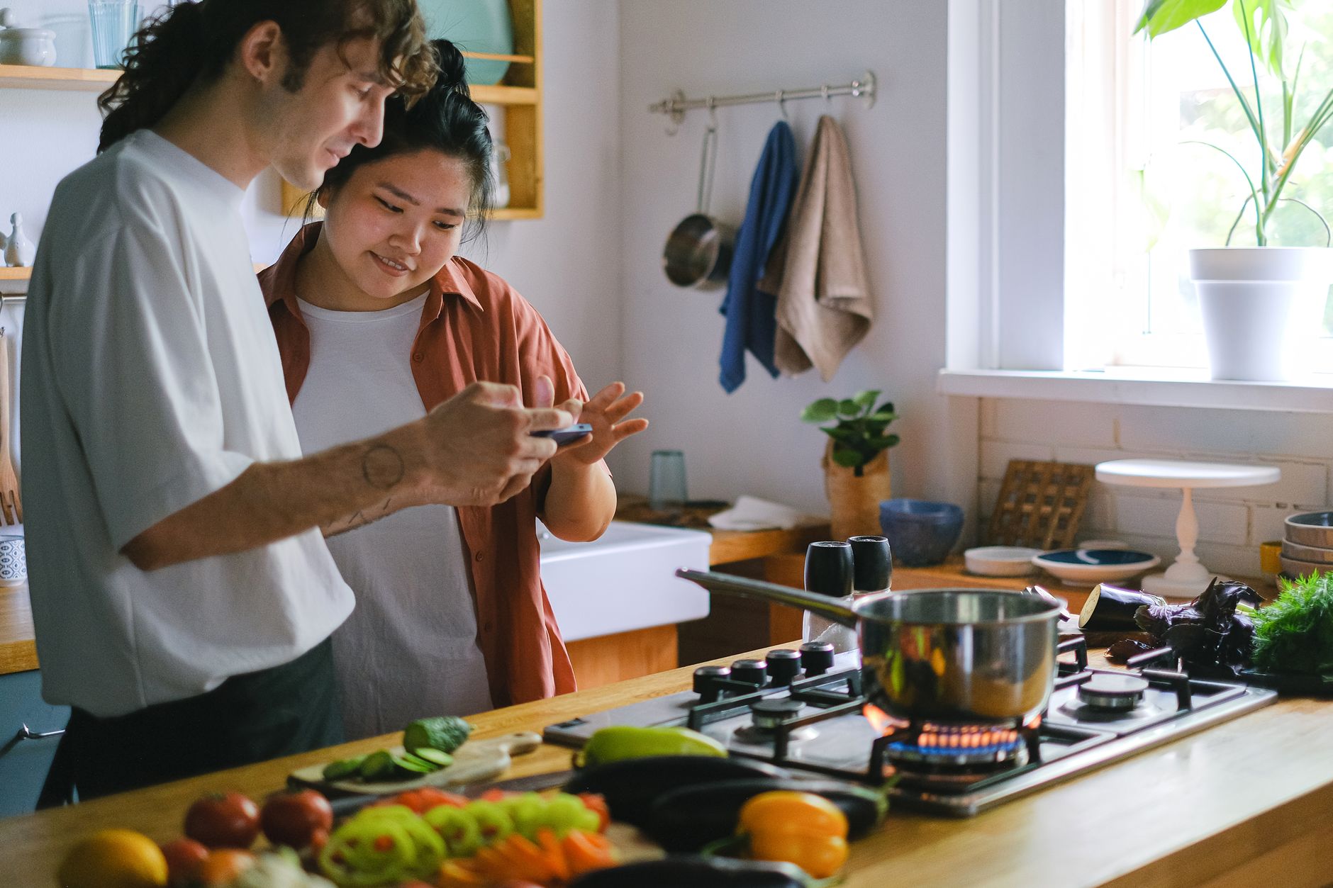 A young couple uses a phone while preparing a meal in their modern kitchen with fresh vegetables.