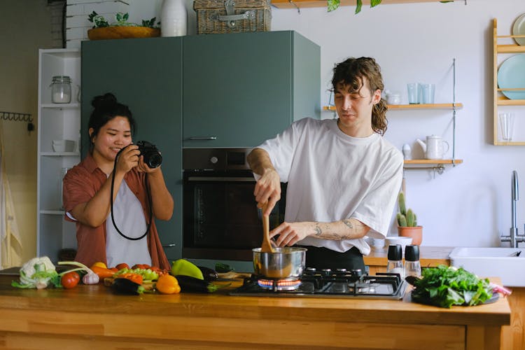 Woman Taking A Picture Of A Man Cooking In A Kitchen