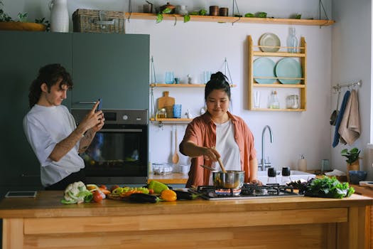 Asian woman cooking while man takes pictures in a stylish kitchen setting.