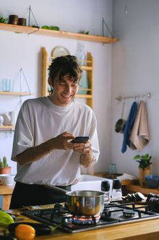 Smiling man using a smartphone while cooking on a stove in a cozy kitchen setting.