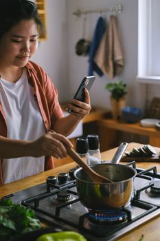 Young woman multitasking by cooking and using her smartphone in a cozy kitchen.