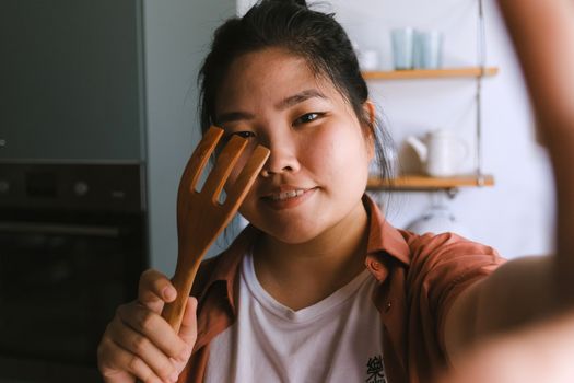 Smiling young woman posing with a wooden kitchen utensil in a modern kitchen setting.