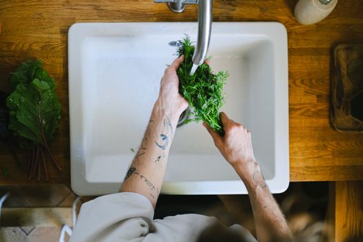 Tattooed hands washing fresh herbs in a kitchen sink, highlighting daily culinary routines.