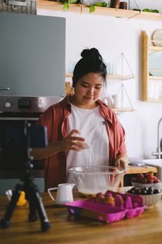 Young woman making a cooking tutorial video in a modern kitchen using a phone and tripod.
