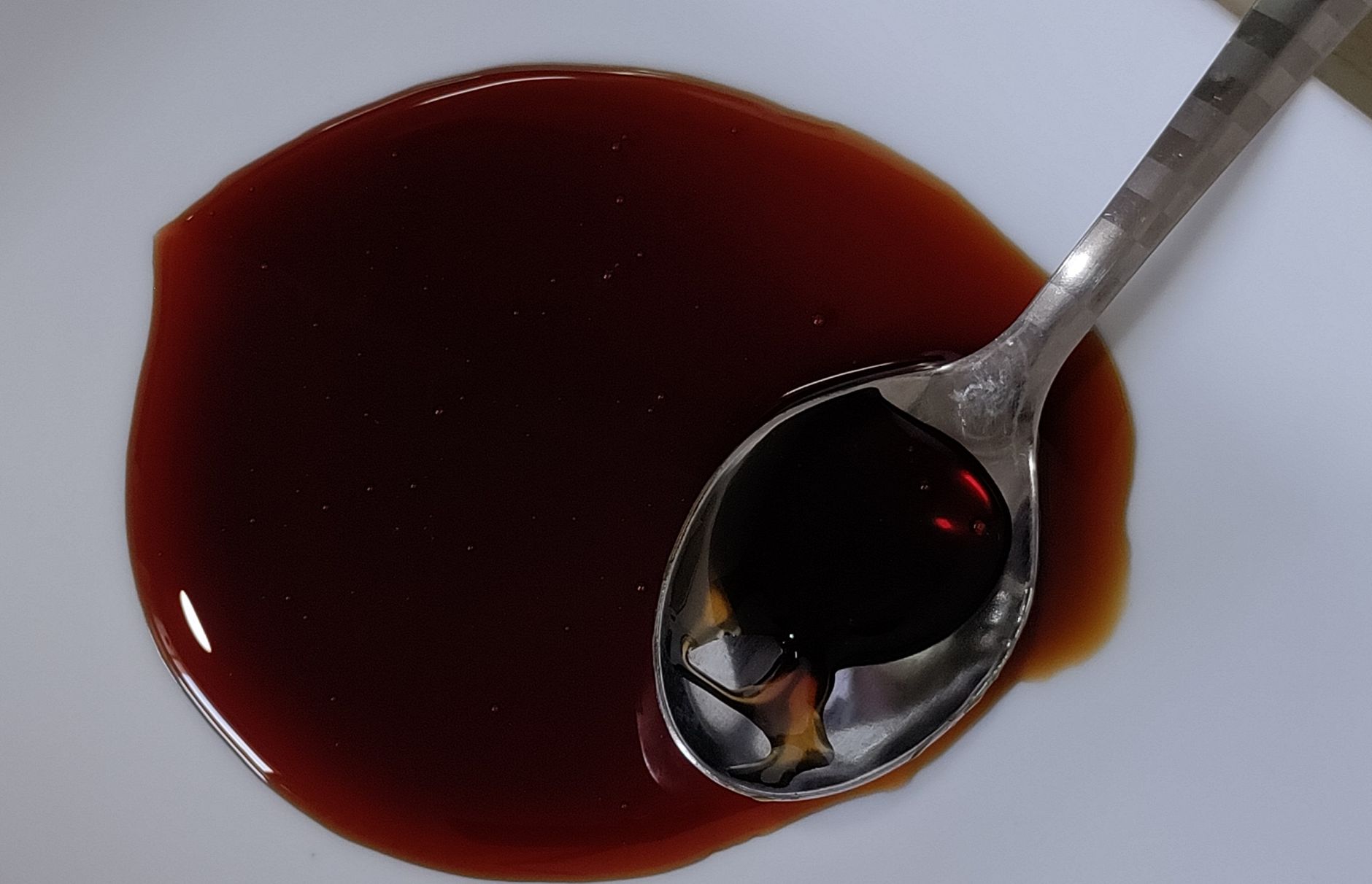 A close-up image of a spoon holding dark soy sauce on a white ceramic plate.