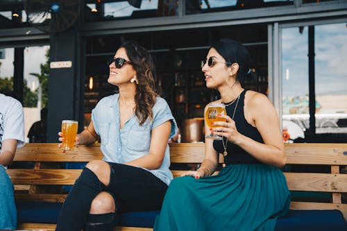 Woman in Black and Blue Sleeveless Dress Holding Glass of Beer Sitting on Bench