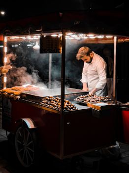 A street vendor prepares roasted chestnuts at night under warm lights and steam.