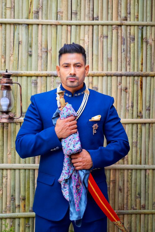 Free Portrait of a South Asian man in a blue Jodhpuri suit holding a sword against a bamboo backdrop. Stock Photo
