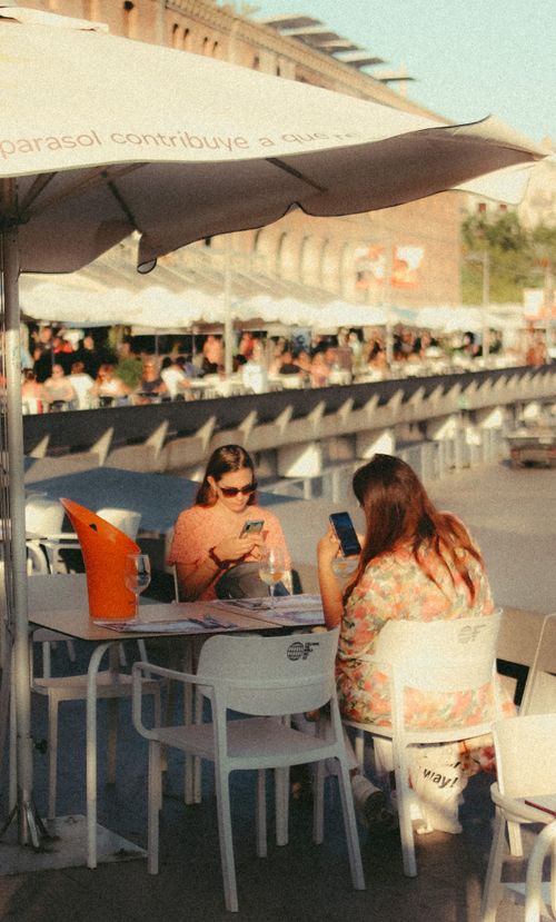 Free Two women sit at an outdoor cafe, absorbed in their smartphones on a sunny day. Stock Photo