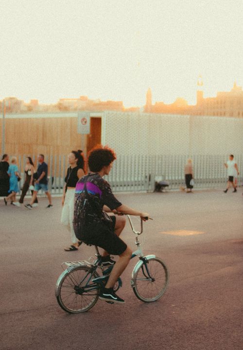 Free A person rides a bicycle on a city street at sunset, surrounded by people walking and modern buildings. Stock Photo