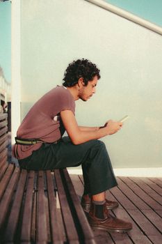 A young man sits on a wooden bench outdoors, engrossed in his smartphone.