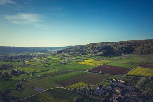 Free Aerial view of lush green farm fields with a distant village and clear blue sky. Stock Photo