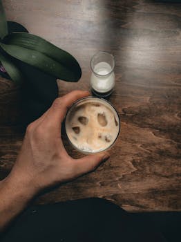 Aerial view of hand holding iced coffee with milk on wooden surface.