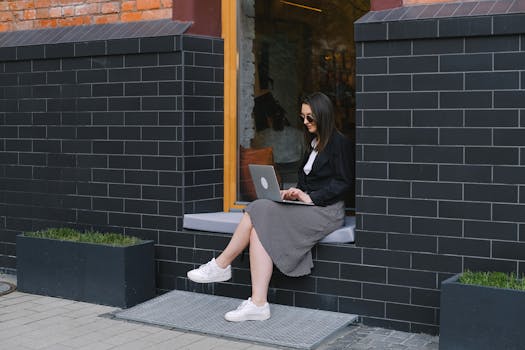 Woman in sunglasses working on laptop outdoors, combining style and productivity in a modern urban setting.